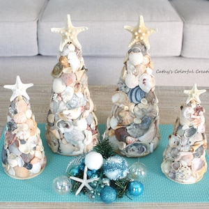 May include: Four seashell Christmas trees of varying sizes, each topped with a starfish. The trees are decorated with a variety of seashells in neutral tones. Decorative blue and silver ornaments and a starfish are arranged in front of the trees.