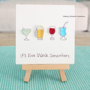 May include: A small white canvas with four colorful drink illustrations: a martini, beer, red wine, and a blue cocktail. The phrase "It's Five O'clock Somewhere" is written below the drinks. The canvas is displayed on a small wooden easel.