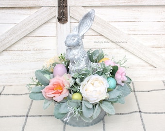 Peut inclure: Une figurine de lapin en argent est placée sur un pot en métal galvanisé rempli d'un arrangement floral de printemps. L'arrangement comprend des fleurs roses, blanches et jaunes, ainsi que du feuillage vert et des œufs de Pâques aux couleurs pastel.