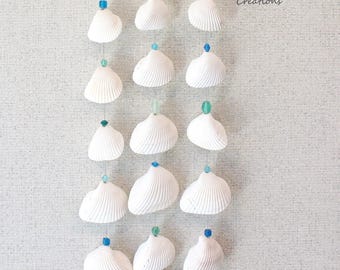 Könnte beinhalten: Ein Windspiel aus weißen Muscheln mit blauen und grünen Glasperlen, die an einem Treibholzast hängen. Das Windspiel trägt den Text "Cathy's Colorful Creations".