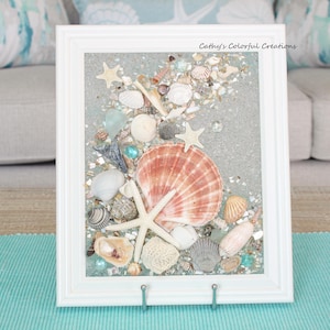 May include: A framed artwork featuring a collection of seashells, starfish, and sea glass. The artwork is set against a light blue background and framed in a white rectangular frame. The text "Cathy's Colorful Creations" is visible in the upper right corner.