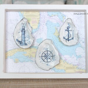 May include: A white rectangular frame containing a nautical map background and three oyster shells. The shells feature blue ink illustrations of a lighthouse, an anchor, and a compass rose. The text Cathy's Colorful Creations is visible.