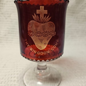 May include: A ruby red glass votive candle holder with a clear glass stem. The holder features a gold-colored Sacred Heart design with a cross and the words "THY KINGDOM COME". The rim has a scalloped edge.