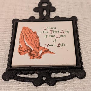 May include: A black cast iron trivet with a white ceramic tile insert. The tile has a picture of two hands clasped in prayer and the text "Today is the First Day of the Rest of Your Life."