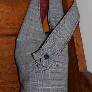May include: A gray and black plaid fabric handbag with a ruffled trim and a black button. The bag has a burgundy lining and a long strap.