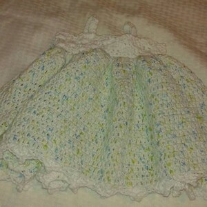 Puede incluir: Un vestido de beb&eacute; blanco de ganchillo con un patr&oacute;n moteado verde y azul. El vestido tiene un borde de encaje festoneado.