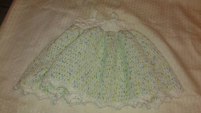 Puede incluir: Un vestido de beb&eacute; blanco de ganchillo con un patr&oacute;n moteado verde y azul. El vestido tiene un borde de encaje festoneado.