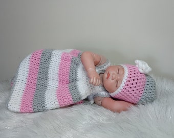 May include: A pink, white, and gray striped crocheted baby sleeping bag with a matching crocheted hat. The sleeping bag is laid out on a white fluffy surface.