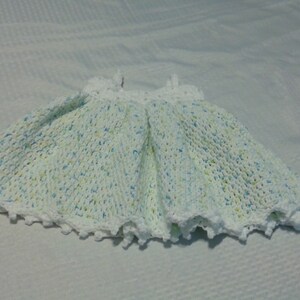 Puede incluir: Un vestido de beb&eacute; de crochet blanco con un patr&oacute;n moteado azul claro y verde. El vestido tiene un borde festoneado y es perfecto para una ocasi&oacute;n especial.