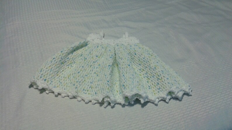 Puede incluir: Un vestido de beb&eacute; de crochet blanco con un patr&oacute;n moteado azul claro y verde. El vestido tiene un borde festoneado y es perfecto para una ocasi&oacute;n especial.