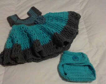 May include: A crocheted baby dress and diaper cover set in teal and gray. The dress has a ruffled hem and a flower detail. The diaper cover has a button closure.
