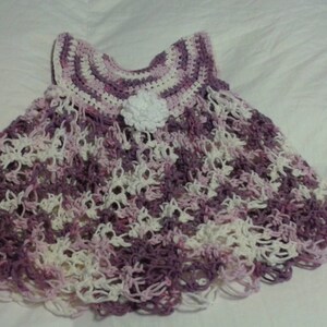 May include: A crocheted baby dress in shades of purple and white with a white flower embellishment.