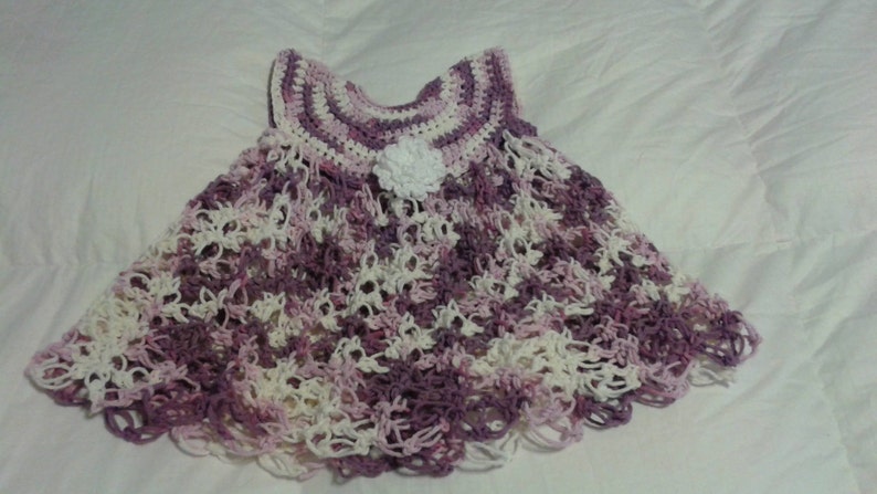May include: A crocheted baby dress in shades of purple and white with a white flower embellishment.