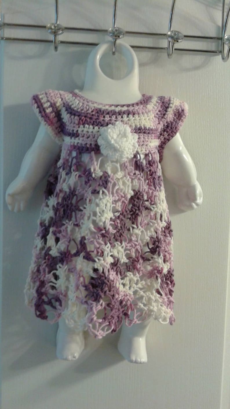 May include: A purple and white crocheted dress with a flower detail, displayed on a white doll form.