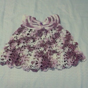 May include: A crocheted baby dress in shades of purple and white. The dress has a scalloped hem and a delicate lace pattern.