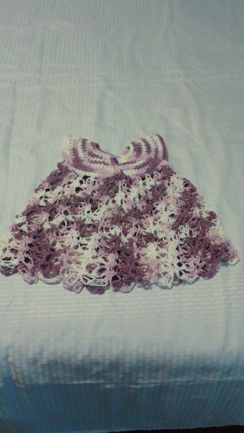 May include: A crocheted baby dress in shades of purple and white. The dress has a scalloped hem and a delicate lace pattern.