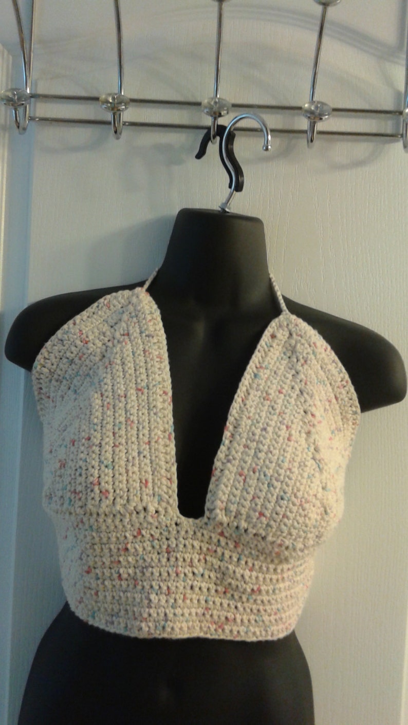 Puede incluir: Un top de crochet blanco con tirantes y un patr&oacute;n moteado de rosa, azul y blanco. El top tiene un escote en V profundo y es corto.