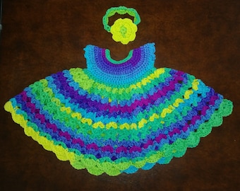 May include: A colorful crocheted baby dress with a matching headband. The dress is made in shades of green, blue, purple, and yellow. The headband has a flower design.