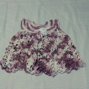 May include: A crocheted baby dress in shades of purple and white. The dress has a scalloped hem and a small white flower embellishment.