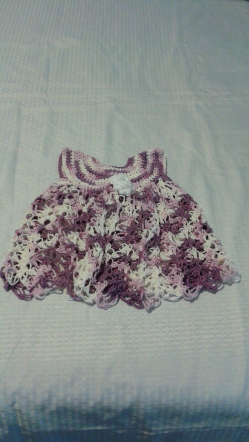 May include: A crocheted baby dress in shades of purple and white. The dress has a scalloped hem and a small white flower embellishment.