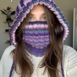 May include: A knitted hood in shades of purple, blue, and pink, with a matching face covering. The hood features a striped pattern and long braided ties. The person is wearing a white sweater.