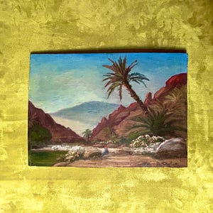 Puede incluir: Pintura al óleo de un oasis en el desierto bajo un cielo azul. La obra presenta una palmera inclinada, formaciones rocosas rojas y una masa de agua. Flores blancas y pequeñas figuras son visibles en primer plano. La pintura está firmada abajo a la derecha.