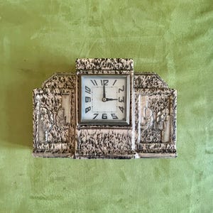 May include: An antique Art Deco clock with a square face and a decorative, textured frame. The clock face displays the time as 2:59. The frame has a speckled, cream and black finish, with floral relief details on the sides.