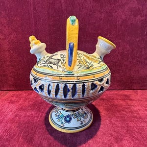 Könnte beinhalten: Handbemalter Keramikkrug mit gelbem Griff und Ausguss. Der Krug hat ein detailliertes Design mit blauen, gelben und orangen Akzenten und einem floralen Sockel. Der Körper hat dekorative Aussparungen.