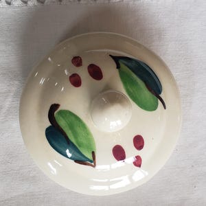 May include: Cream-colored ceramic lid with a central knob. The lid is decorated with green leaves, dark blue accents, and small red berry clusters. A decorative piece for kitchen or dining.