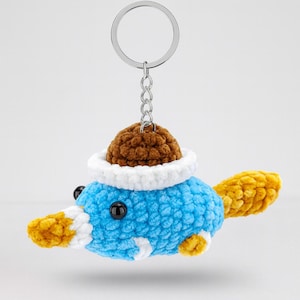 Puede incluir: Llavero de ganchillo hecho a mano con forma de ornitorrinco azul con sombrero marrón y cola y pico amarillos. El llavero tiene un anillo y una cadena de metal plateado. El ornitorrinco tiene ojos negros y detalles blancos.