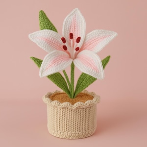Pode incluir: Uma flor de lírio de crochê em um vaso bege. A flor tem pétalas brancas com detalhes em rosa e manchas vermelhas. Folhas verdes cercam a flor. O vaso é bege com uma borda ondulada, e o interior é preenchido com material bege.