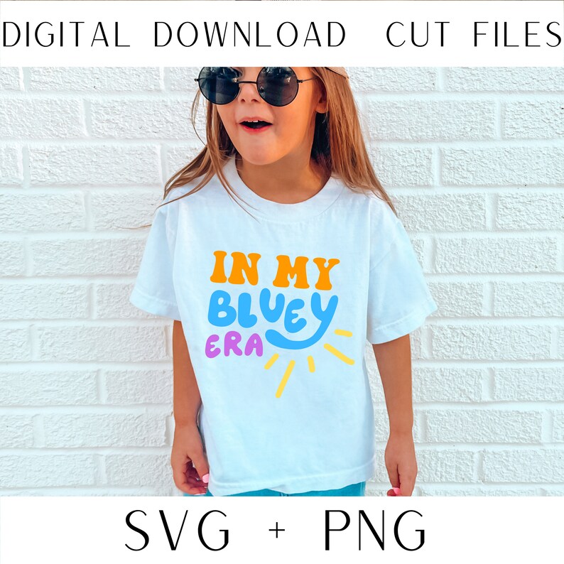 May include: A white t-shirt with the text "In My Bluey Era" in colorful, stylized lettering. The text is surrounded by a yellow sun with rays.