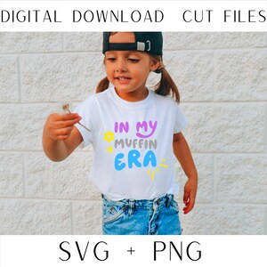 May include: A white t-shirt with the text "In My Muffin Era" in colorful, bubble-like letters. The shirt is being worn by a child with a black baseball cap.