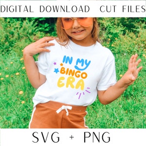 May include: A white t-shirt with the text "In My Bingo Era" in colorful, playful lettering. The shirt is being worn by a child in a grassy field.