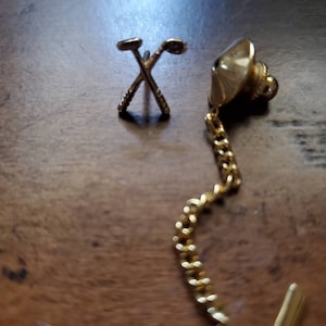 May include: A gold-toned golf club lapel pin with a chain attached. The pin features two crossed golf clubs and a small, round, textured design.