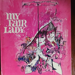 May include: A pink book cover with the title "My Fair Lady" in white lettering. The cover features a colorful illustration of a couple under an umbrella, surrounded by other people.