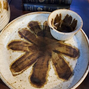 May include: A white ceramic serving dish with a brown flower design. The dish is slightly raised around the edges and has a small, white ceramic bowl in the center.
