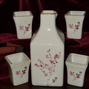 May include: A set of five white ceramic sake cups and a matching pitcher with a floral design. The cups and pitcher have a gold rim and feature pink flowers on a white background.