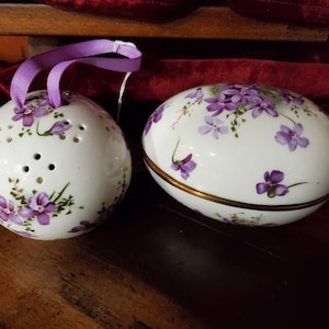 May include: Two white ceramic egg-shaped ornaments with purple floral designs. One ornament has a lid and the other has holes for air circulation.