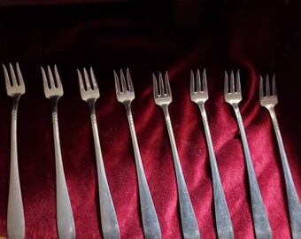 May include: Eight silver dessert forks with ornate handles laid out on a red velvet surface.