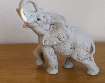 Può includere: Statuetta di elefante grigio chiaro con proboscide sollevata e zanne d'avorio. L'elefante è in posizione di camminata, con dettagli strutturati sulla sua pelle. La statuetta è su una superficie di legno.