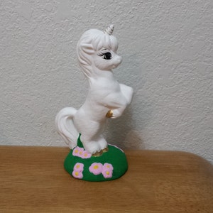 May include: A white ceramic unicorn figurine standing on its hind legs, with a golden horn and hooves. The unicorn is on a green base with pink flower accents. The figurine is displayed on a wooden surface.