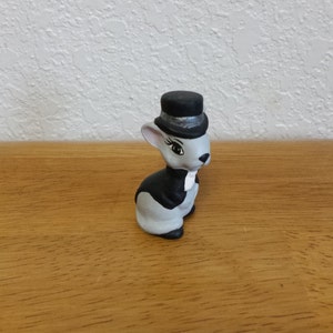May include: A ceramic figurine of a rabbit wearing a black top hat and tuxedo. The rabbit is gray with black accents and a white bow tie. The figurine is sitting on a wooden surface.