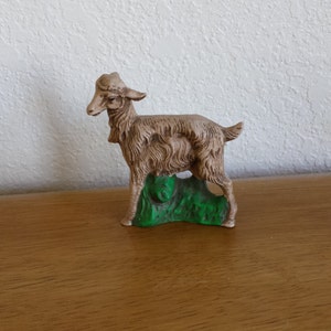 以下が含まれることがあります： 緑色の台座の上に立っている、毛並みの詳細なタン色のヤギの置物。 ヤギは小さなひげがあり、立っているポーズをとっています。 この置物は、セラミックまたは同様の素材で作られている可能性があります。