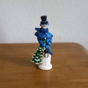 Puede incluir: Una figura de muñeco de nieve azul y blanco que sostiene un pequeño árbol de Navidad verde. El muñeco de nieve lleva un sombrero y una bufanda azules.