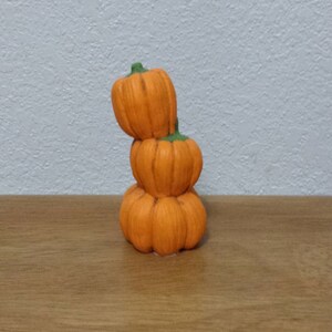 Könnte beinhalten: Ein Stapel aus drei kleinen, orangefarbenen Keramikkürbissen mit grünen Stielen. Die Kürbisse sind übereinander gestapelt, wobei der größte Kürbis unten und der kleinste Kürbis oben liegt.