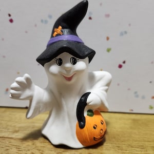 Op de afbeelding: Een wit spookfiguurtje met een zwarte heksenhoed met een paarse band en een oranje kruis. Het spookje houdt een kleine oranje pompoen vast met een pompoengezicht.