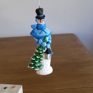May include: A blue and white snowman ornament holding a small green tree and a lantern. The snowman is wearing a blue hat and scarf.