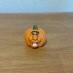 May include: A small orange pumpkin with a green stem and a painted face. The pumpkin has a surprised expression with wide eyes and an open mouth.