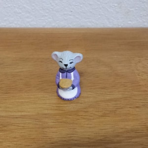 以下が含まれることがあります： 紫色のドレスを着てパイを持っているネズミの小さなセラミック製フィギュア。ネズミは白いエプロンを着ていて、笑顔をしています。